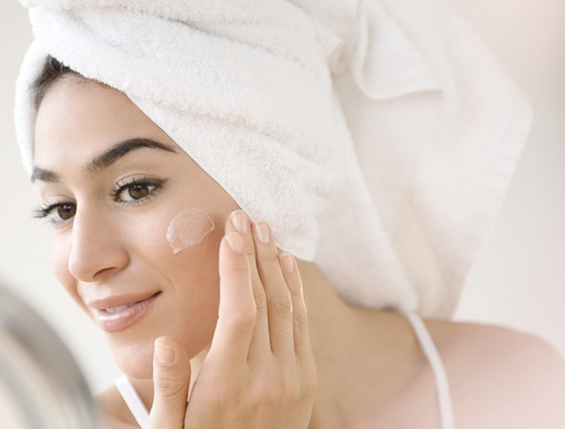 Woman with hair in towel rubbing lotion on face