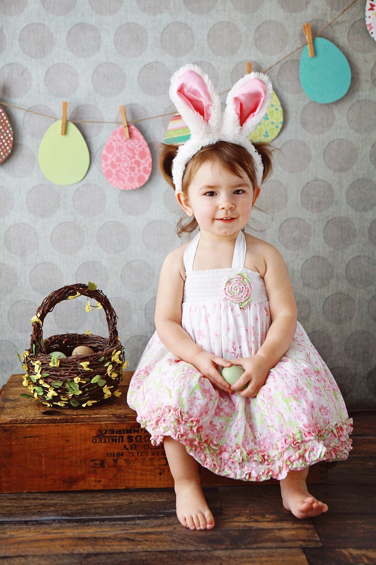 A beautiful little girl smiles at the camera as she wears a pair of bunny ears. A basket filled with easter eggs is beside her. A decorative garland of colourful easter eggs hangs behind her.