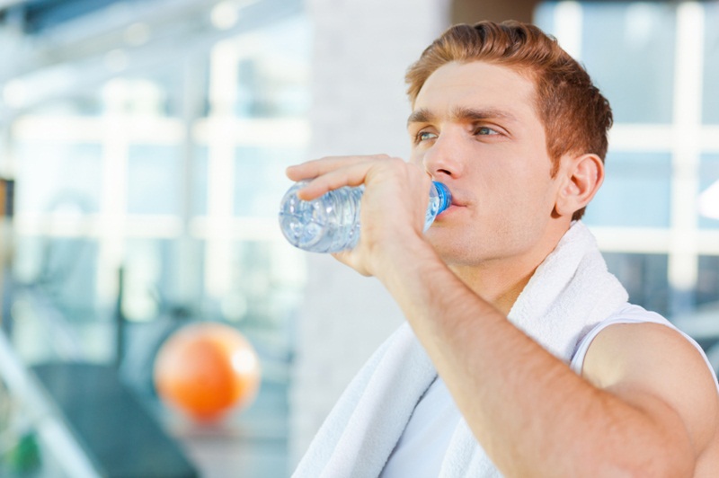 Refreshing after work out. Tired young man carrying towel on shoulders and drinking water while standing in gym