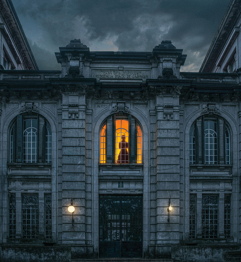 Hình ảnh tăm tối của bệnh viện tâm thần hoang phế với ánh đèn bí ẩn bên trong tòa nhà, cùng một bóng hình u ám tựa như một linh hồn lẩn khuất.